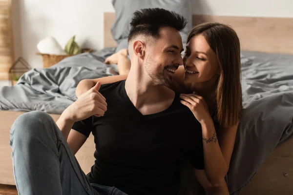 Hombre feliz en camiseta negra mirando a la novia joven abrazándolo mientras está acostado en la cama - foto de stock