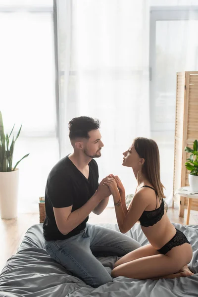 Молода жінка в сексуальній білизні і чоловік в чорній футболці тримає руки і дивиться один на одного в спальні — стокове фото