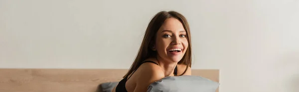 Щаслива спокуслива жінка дивиться на камеру, обіймаючи сіру подушку в спальні, банер — стокове фото