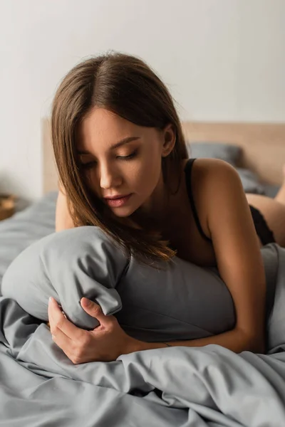 Joven sensual mujer abrazando almohada mientras está acostado en la cama en casa - foto de stock