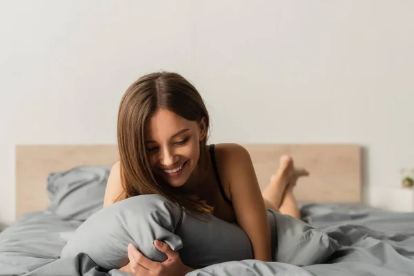 Young and sensual woman smiling while lying on grey bedding at home — Stock Photo