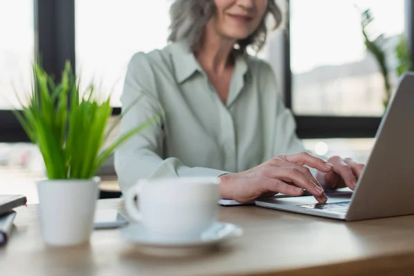 Vista recortada de la mujer de negocios borrosa usando el ordenador portátil cerca del café y la planta en la oficina - foto de stock