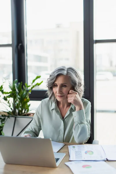 Зрелая деловая женщина с ноутбуком рядом с документами на столе в офисе — стоковое фото