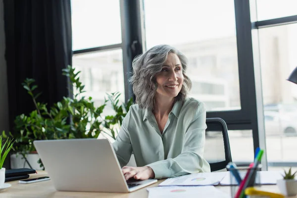 Sonriente mujer de negocios madura utilizando el ordenador portátil cerca de los papeles en la oficina - foto de stock