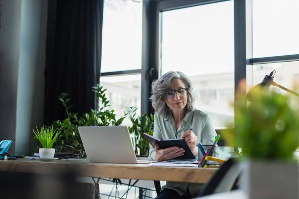 Reife Geschäftsfrau mit Brille schreibt auf Notizbuch in der Nähe von Laptop und Fabrik im Büro — Stockfoto