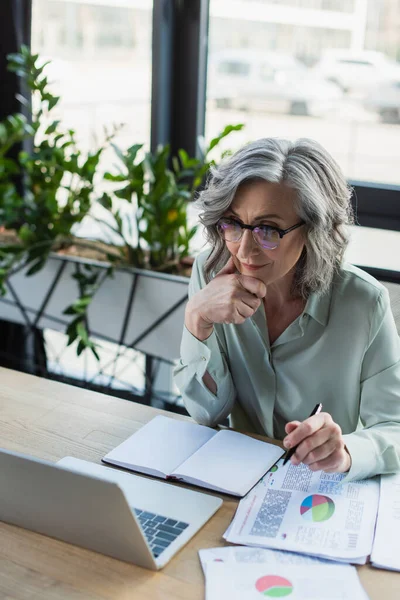 Empresária de cabelos grisalhos segurando caneta perto de papelada, caderno e laptop no escritório — Fotografia de Stock