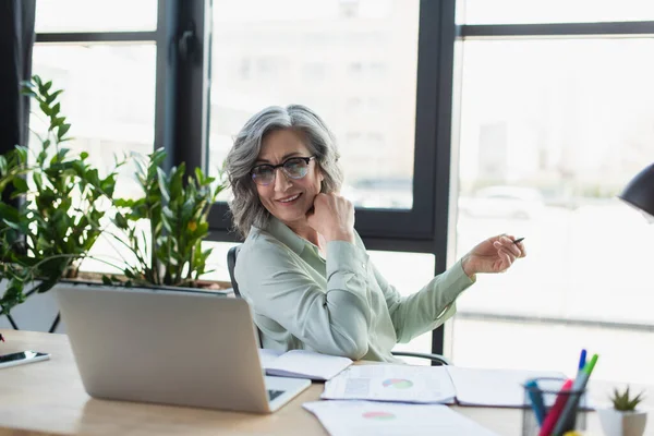 Cheerful businesswoman holding pen near documents and devices in office — Stock Photo