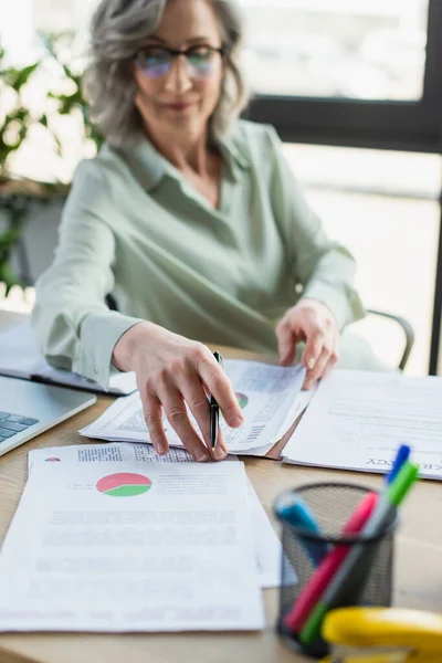 Documents and laptop near blurred businesswoman working in office — Stock Photo