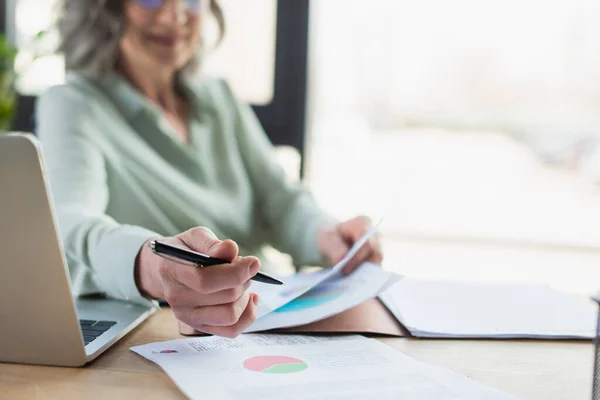 Ausgeschnittene Ansicht einer verschwommenen Geschäftsfrau mit Stift in der Nähe von Papieren und Laptop im Büro — Stockfoto