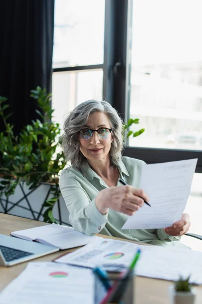 Mature businesswoman holding documents near blurred laptop and notebook in office — Stock Photo