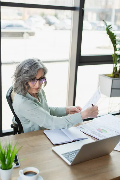 Middle aged businesswoman holding document near devices and coffee on table in office — Stock Photo