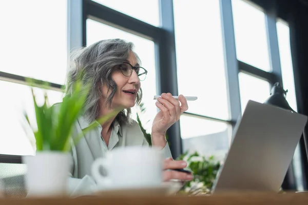 Femme d'affaires mature enregistrant un message vocal sur un téléphone portable près d'un ordinateur portable et une tasse de café floue au bureau — Photo de stock