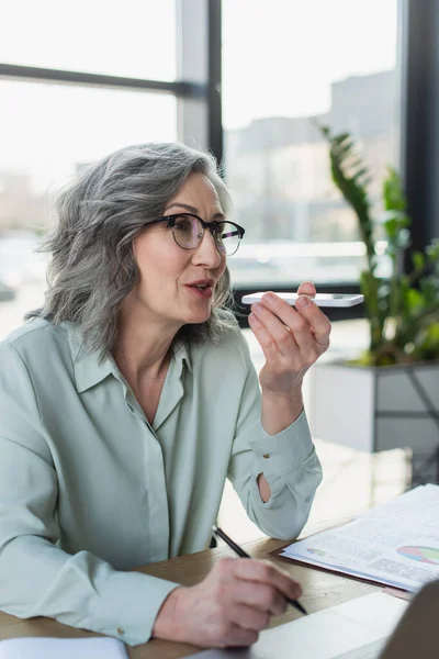Grauhaarige Geschäftsfrau nimmt Sprachnachricht auf Handy auf und hält Stift in der Nähe von Laptop im Büro — Stockfoto