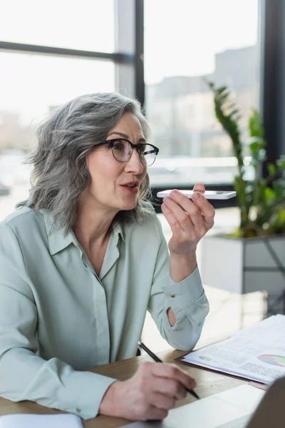 Mujer de negocios de pelo gris grabando mensaje de voz en el teléfono celular y sosteniendo la pluma cerca de la computadora portátil en la oficina - foto de stock