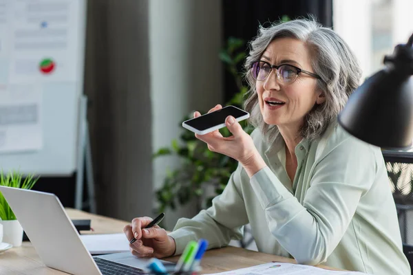 Madura mujer de negocios grabación de mensajes de voz en el teléfono inteligente y el uso de ordenador portátil en la oficina - foto de stock