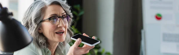 Grauhaarige Geschäftsfrau mit Brille, die Sprachnachricht auf dem Smartphone aufzeichnet, neben verschwommener Lampe im Büro, Banner — Stockfoto