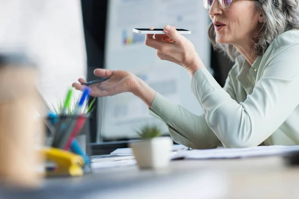 Cropped view of middle aged businesswoman recording voice message on mobile phone near papers in office — Stock Photo