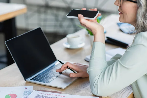 Vista recortada de una mujer de negocios de cabello gris sosteniendo un teléfono inteligente con pantalla en blanco y utilizando un ordenador portátil cerca de documentos con gráficos en la oficina — Stock Photo