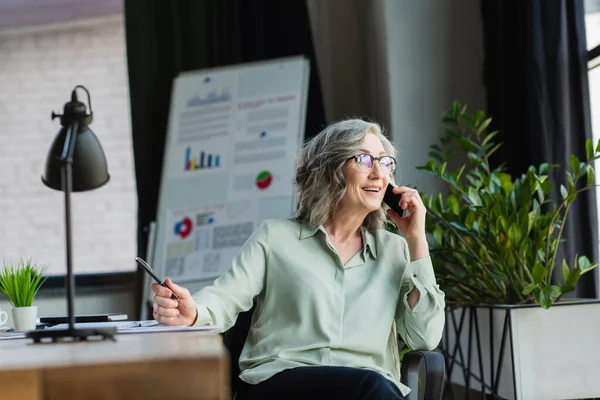 Mujer de negocios sonriente sosteniendo pluma mientras toma el teléfono inteligente cerca de las plantas y el rotafolio borroso en la oficina - foto de stock