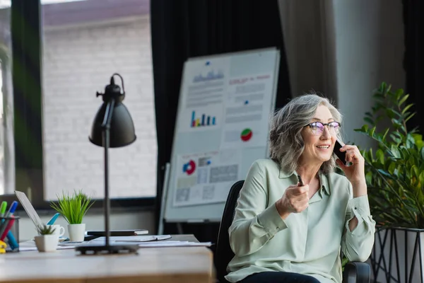 Middle aged businesswoman talking on cellphone near working table in office — Stock Photo