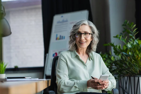 Mature businesswoman smiling at camera while holding pen and smartphone in office — Stock Photo