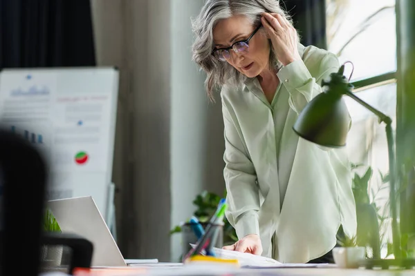 Grauhaarige Geschäftsfrau mit Brille schaut sich Dokumente in der Nähe von Laptop im Büro an — Stockfoto