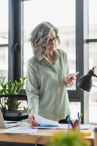 Femme d'affaires d'âge moyen utilisant un téléphone portable et tenant un document près de la papeterie et de la lampe dans le bureau — Photo de stock