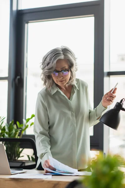 Femme d'affaires mature en lunettes tenant smartphone près de documents au bureau — Photo de stock