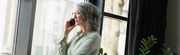 Femme d'affaires aux cheveux gris parlant sur smartphone près de la fenêtre dans le bureau, bannière — Photo de stock
