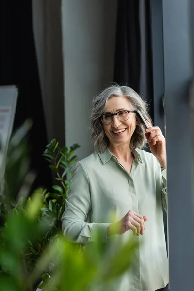 Happy businesswoman in eyeglasses talking on smartphone in office — Stock Photo