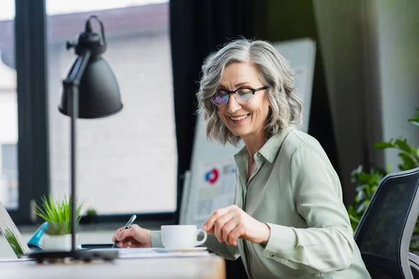 Happy businesswoman in eyeglasses taking coffee cup near laptop and papers — Stock Photo