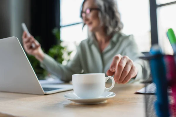 Cup of coffee near blurred businesswoman and laptop in office — Stock Photo