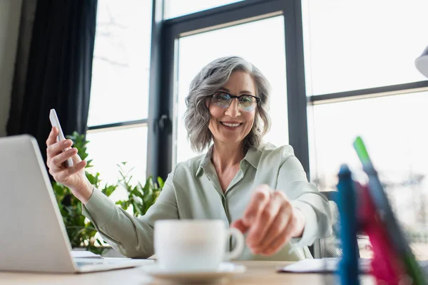 Happy businesswoman holding smartphone and taking cup of coffee in office — Stock Photo