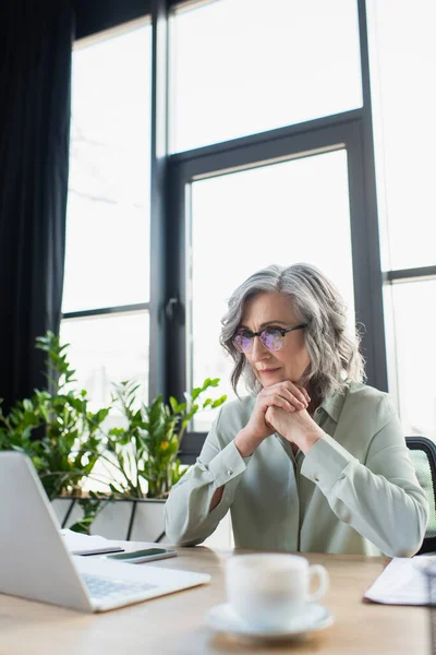 Mature businesswoman looking at laptop near blurred cup of coffee in office — Stock Photo