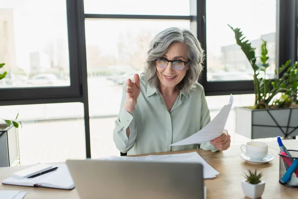 Fröhliche Geschäftsfrau hält Dokument in der Hand und zeigt mit der Hand bei Videoanruf auf Laptop im Büro — Stockfoto