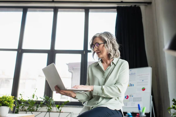 Mature businesswoman in eyeglasses using laptop in office — Stock Photo