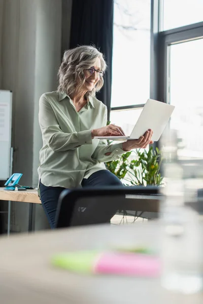 Donna d'affari dai capelli grigi sorridente che utilizza il computer portatile in ufficio — Foto stock