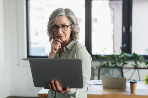 Бізнес-леді середнього віку дивиться на розмитий ноутбук в офісі — стокове фото