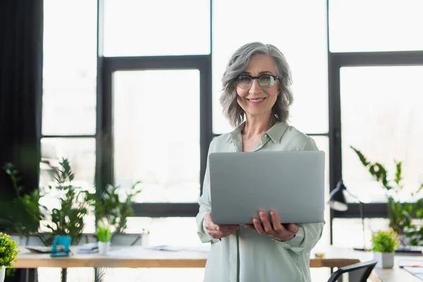 Веселая седовласая деловая женщина в очках держит ноутбук и смотрит в камеру в офисе — стоковое фото