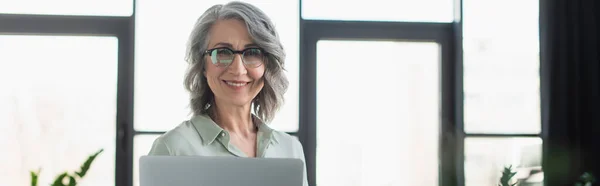 Donna d'affari matura che sorride alla macchina fotografica vicino al computer portatile in ufficio, banner — Foto stock