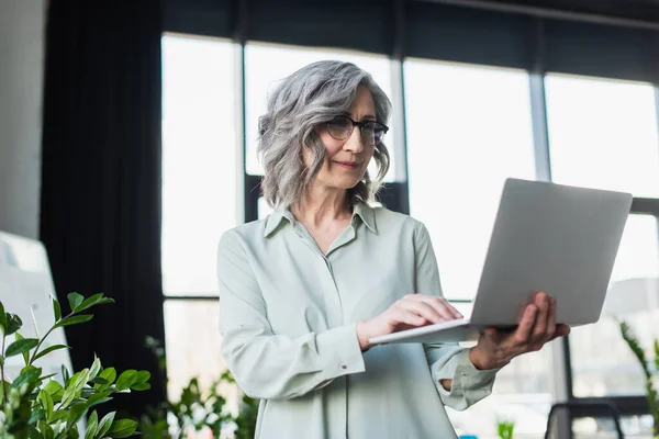 Reife Geschäftsfrau in Brille mit Laptop in der Nähe von Werk im Büro — Stockfoto