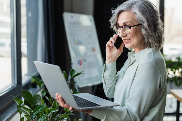 Mujer de negocios de pelo gris hablando por teléfono móvil y sosteniendo el ordenador portátil en la oficina - foto de stock