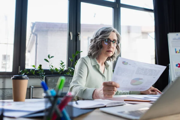 Femme d'affaires aux cheveux gris tenant du papier près du café pour aller et ordinateur portable au bureau — Photo de stock