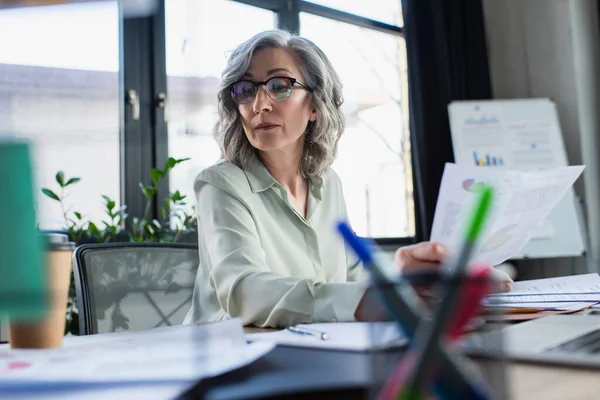 Femme d'affaires aux cheveux gris en lunettes regardant des papiers près d'un ordinateur portable et à emporter boisson au bureau — Photo de stock