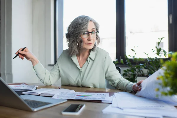 Femme d'affaires tenant un stylo et regardant les papiers près de portable et gadgets dans le bureau — Photo de stock