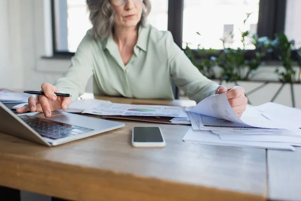 Vista recortada de la mujer de negocios utilizando el ordenador portátil cerca de papeles y teléfonos inteligentes con pantalla en blanco en la oficina - foto de stock