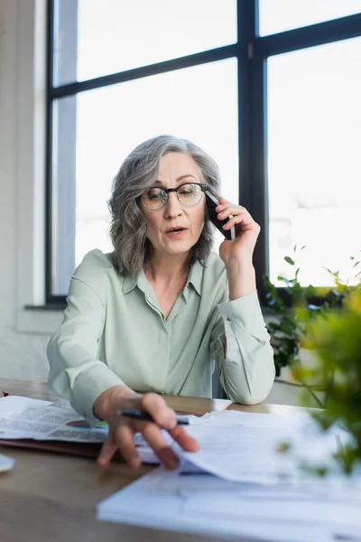 Бізнес-леді розмовляє на мобільному телефоні біля розмитих паперів на столі в офісі — стокове фото