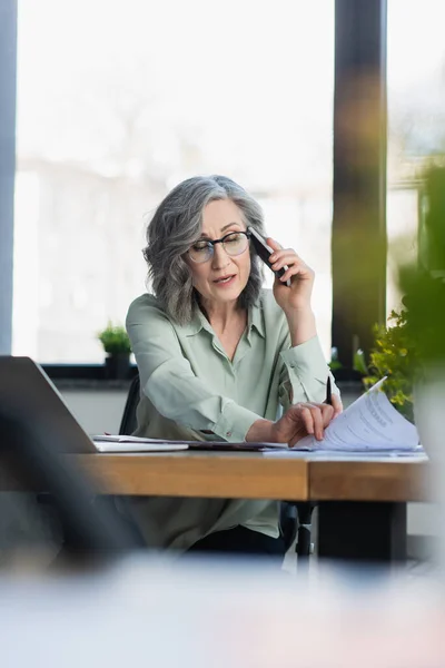 Mujer de negocios de pelo gris mirando documentos mientras habla en el teléfono celular cerca de la mesa de trabajo - foto de stock