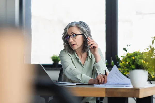 Mature businesswoman talking on smartphone and holding paper near laptop in office — Stock Photo