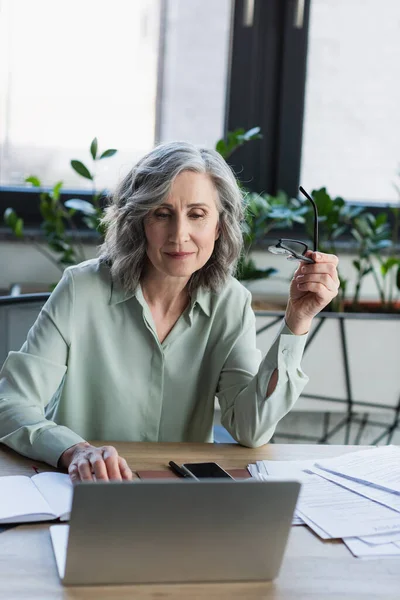Mulher de negócios de meia idade segurando óculos perto de gadgets, papéis e caderno no escritório — Fotografia de Stock